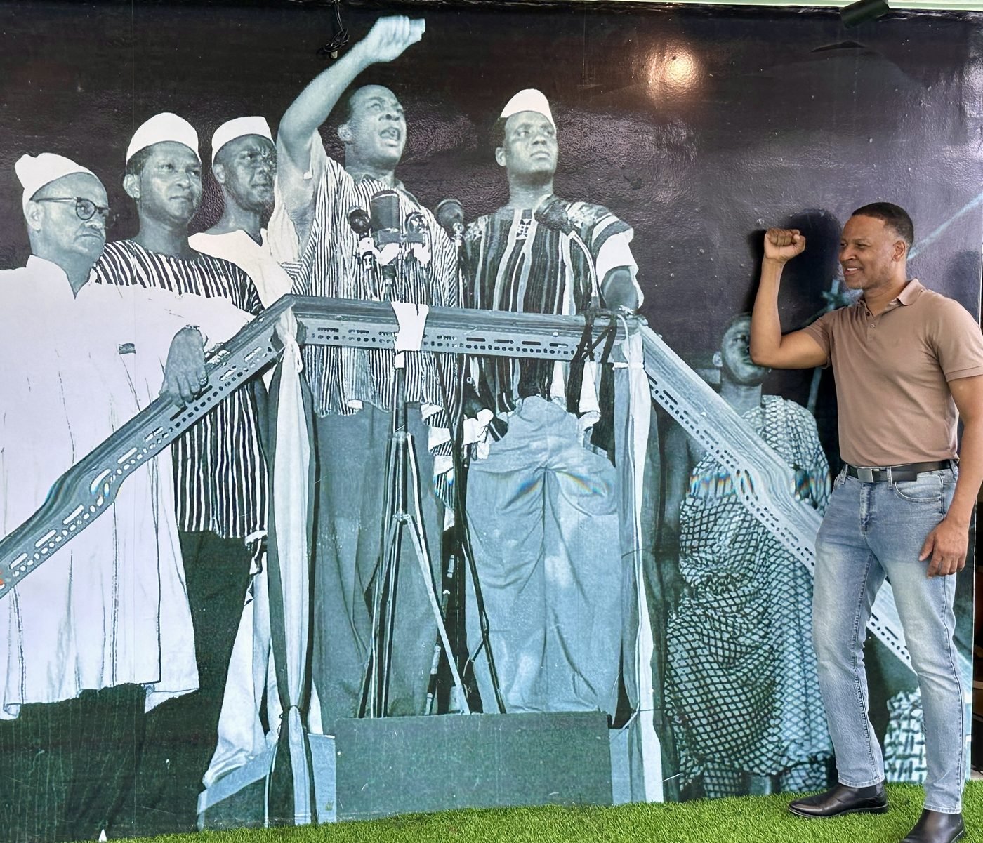 The writer, fist raised, beside a large black-and-white mural of Kwame Nkrumah and his CPP colleagues declaring Ghana's independence in March 1957